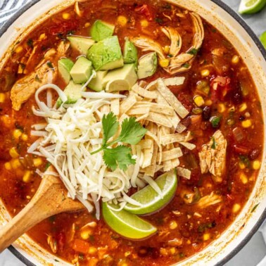 Bird's eye view of chicken tortilla soup topped with cheese, avocado, tortilla strips, lime, and cilantro, served out of a large dutch oven pot with a wooden spoon.