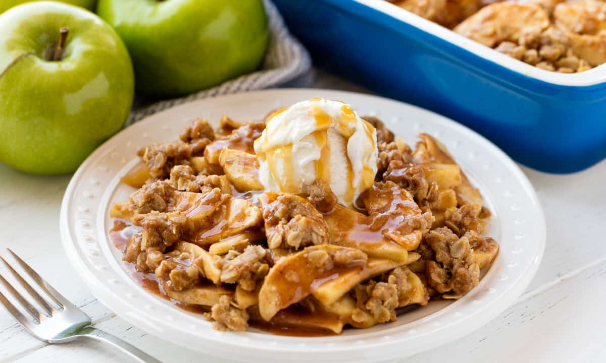 Apple crisp served up on a white plate topped with a scoop of ice cream and caramel sauce.