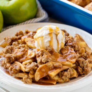 Apple crisp served up on a white plate topped with a scoop of ice cream and caramel sauce.