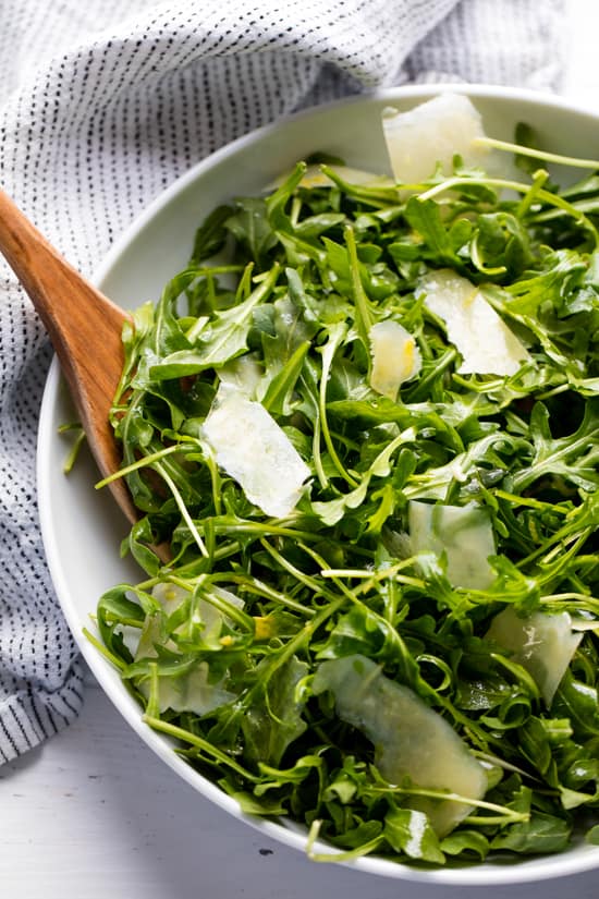 Arugula salad in a white bowl with a wooden spoon in it.