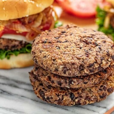 Three Black Bean Burger Patties stacked on a marble slab with a full burger behind complete with bun, lettuce, cheese, onions, and ketchup.