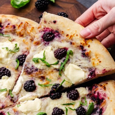 An overhead shot of a slice of blackberry basil ricotta pizza being lifted by a hand away from the rest of the pizza.