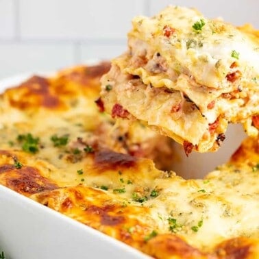 Slice of buffalo chicken lasagna pulled out of a white casserole dish with a bottle of Red Hot buffalo sauce in the background.