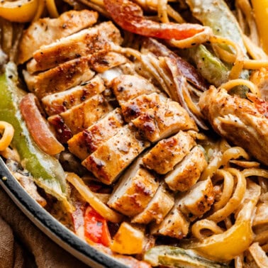 An overhead view of a skillet filled with browned sliced chicken breast, noodles and bell peppers.