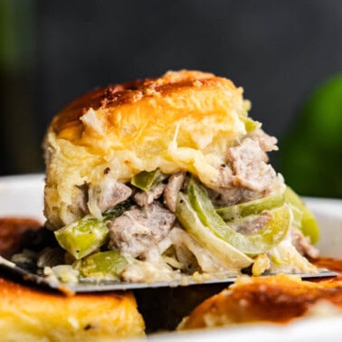 A side shot view of a cheesesteak slider being lifted out of the pan. It's loaded with steak, cheese, bell peppers, and onion.