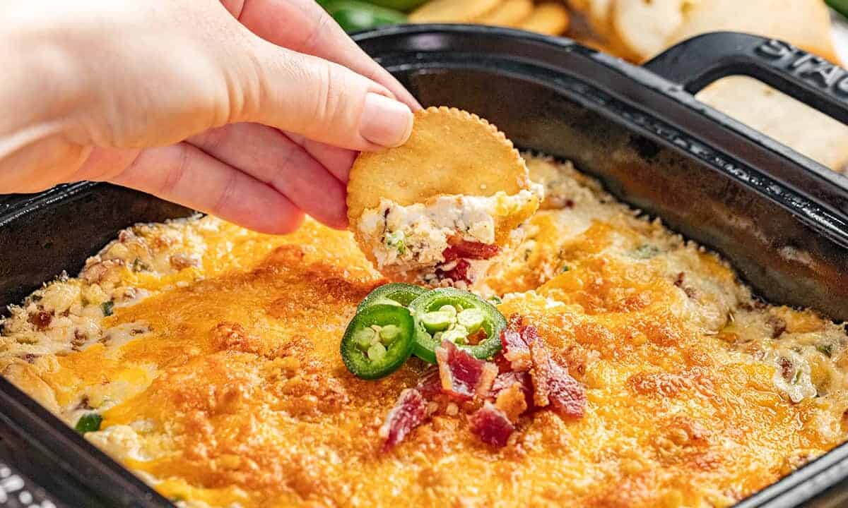 A hand dipping a ritz cracker into cheesy jalapeno popper dip.