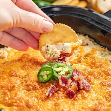 A hand dipping a ritz cracker into cheesy jalapeno popper dip.