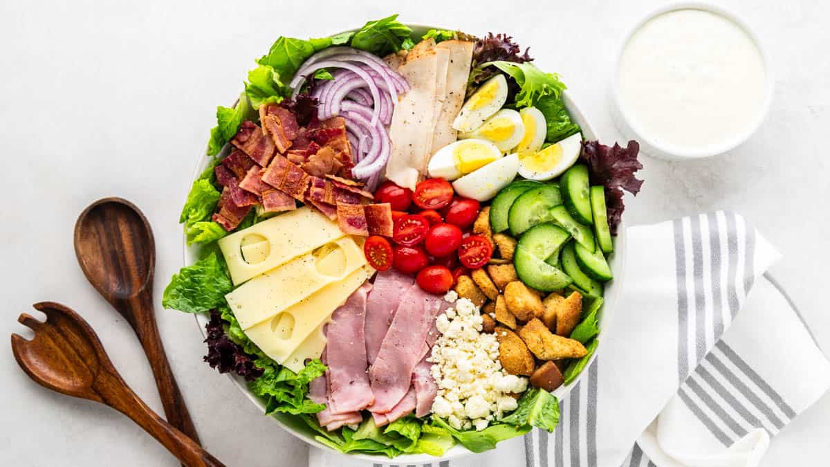 Overhead view of a chef salad with dressing in a small ceramic bowl and wooden serving spoons on the side.