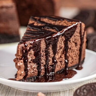 A slice of Chocolate Cheesecake with Oreo Crust, served on a plate and drizzled with chocolate syrup.