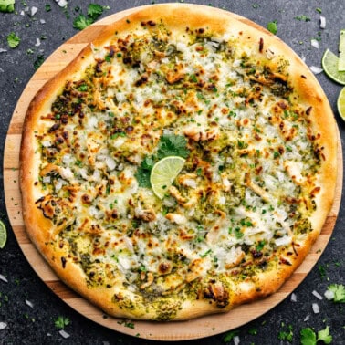 An overhead shot of a full uncut pizza covered in white cheese and green toppings, with fresh limes.