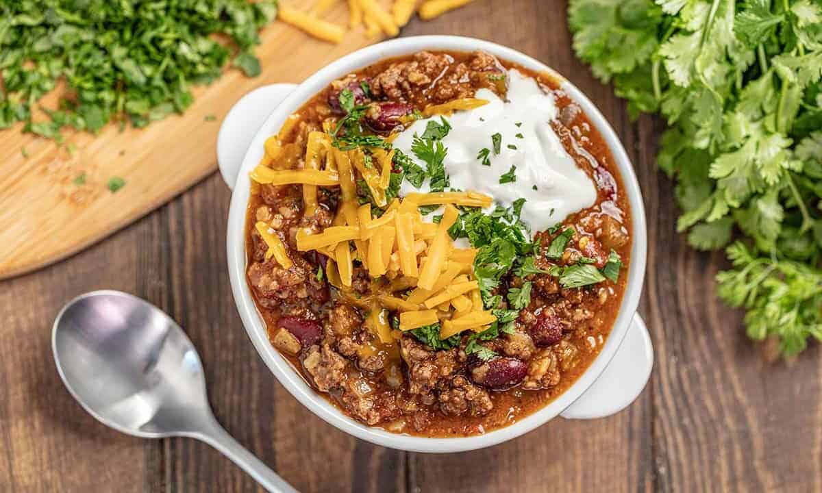 Chili topped with shredded cheese, cilantro and sour cream in a white soup bowl