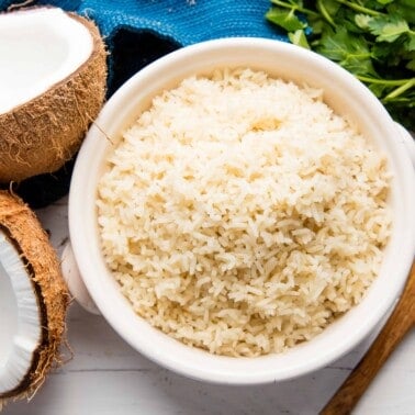 Bird's eye view of Coconut rice in a white bowl by a wooden spoon and two halves of a coconut.