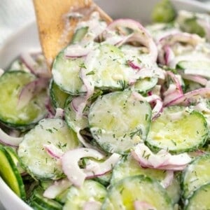 Close up of Creamy Cucumber Salad in a white bowl with a wooden spoon in it.