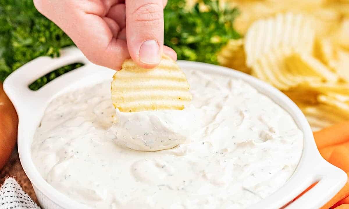 A hand dipping a crinkle cut potato chip into a serving dish filled with creamy French onion dip.