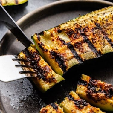 A grilled piece of zucchini is cut with a knife and fork.