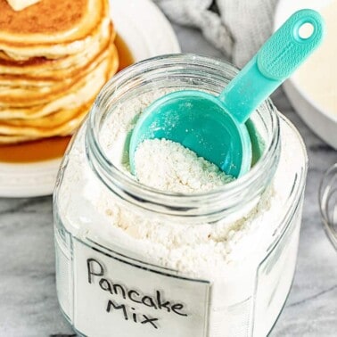 A glass container full of Pancake Mix with a measuring cup in it.