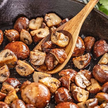 A wooden spoon scooping sautéed mushrooms out of a skillet.