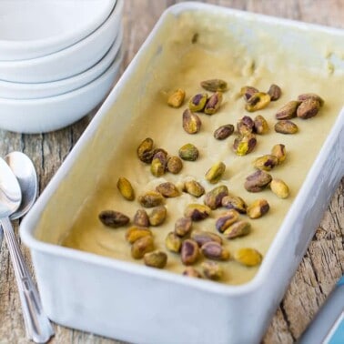 Angled view of Pistachio Gelato in stainless steel bread pan garnished with whole pistachios with stack of white serving bowls on worn wood table top.