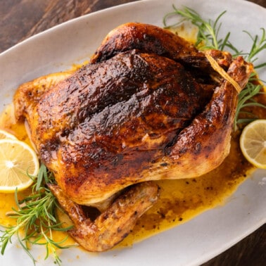 A whole, roasted chicken on a whit serving platter with lemon slices and fresh rosemary sprigs.