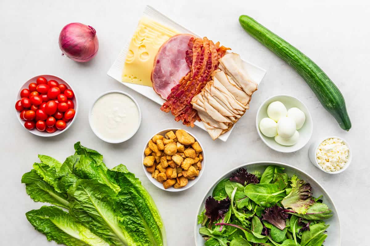 Overhead view of the raw ingredients needed to make a chef salad on a clean, white kitchen counter.