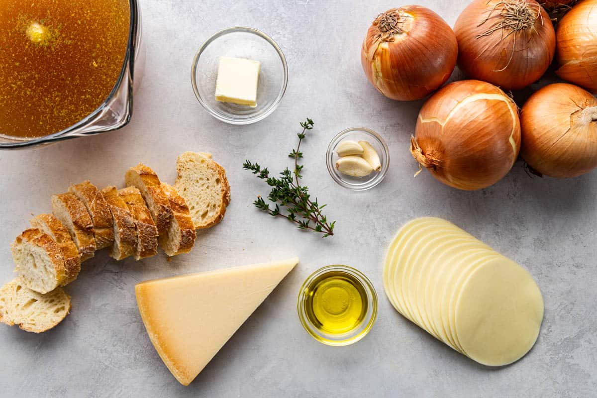 Overhead view of a kitchen counter with yellow onions, beef broth, cheeses, sliced bread, garlic, oil, and fresh herbs.