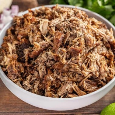 Crockpot carnitas in white bowl, surrounded by limes and corn tortillas