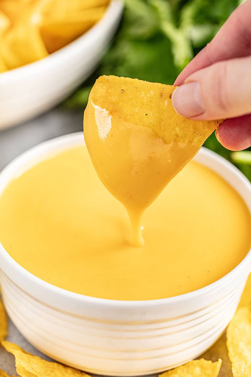 A hand dipping a tortilla chip into a small bowl of cheese sauce