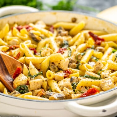 A wooden spoon resting in a pot of Italian chicken and pasta.