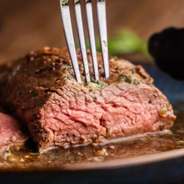 A close up view of a petite tender steak that has already been cut into showing a perfectly pink interior with a fork poking in the top.