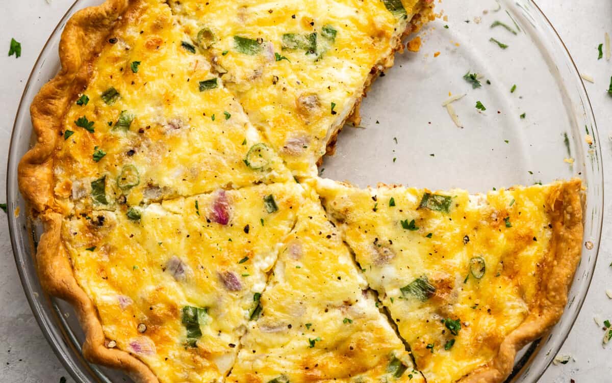 An overhead view of a quiche in a pie plate that is cut and ready to serve with a slice missing.