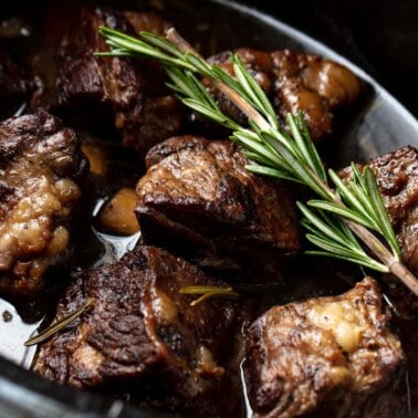 An overhead view of a slow cooker full of beef short ribs with a sprig of rosemary on top.