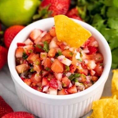 A tortilla chip is dipped into a bowl of strawberry salsa, surrounded by fresh strawberries, lemon, cilantro and tortilla chips