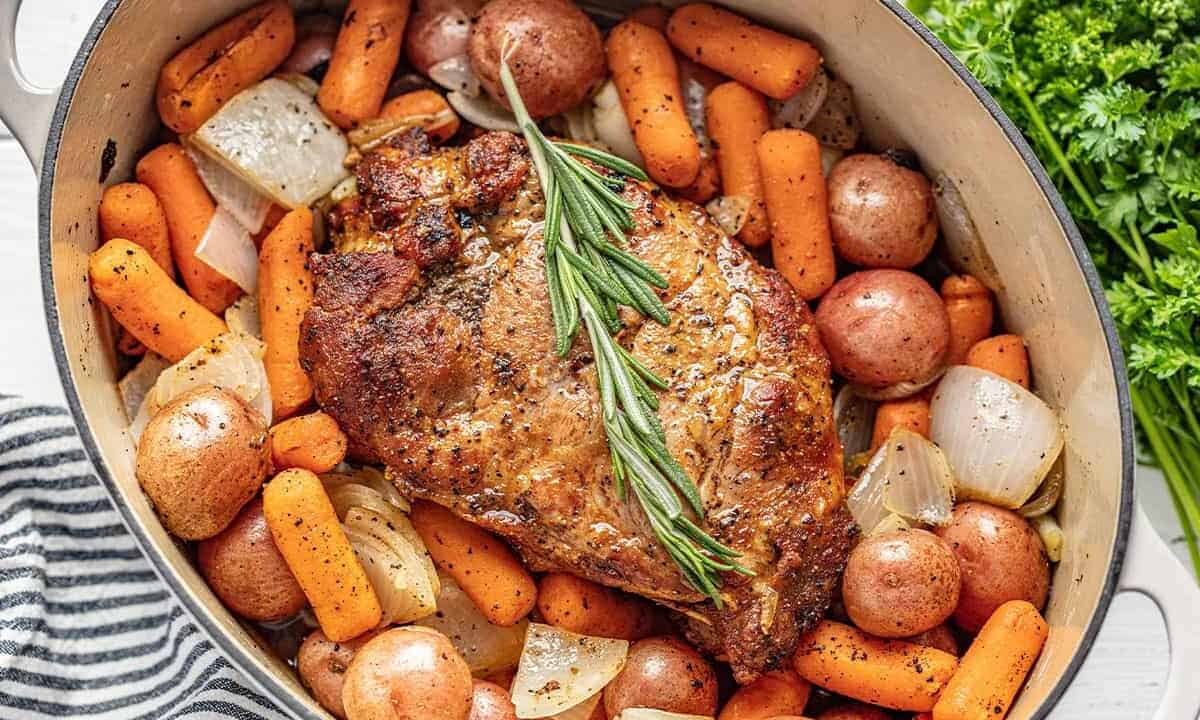 Bird's eye view of Pork Roast garnished with a rosemary sprig, carrots, baby red potatoes, and onion in a cast iron dutch oven.